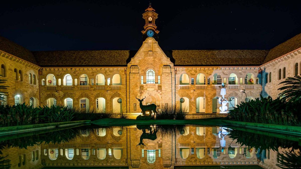 Historisches Gebäude auf dem Hatfield Campus der University of Pretoria bei Nacht beleuchtet. Spiegelung im Teich, Statue eines Springbocks davor.