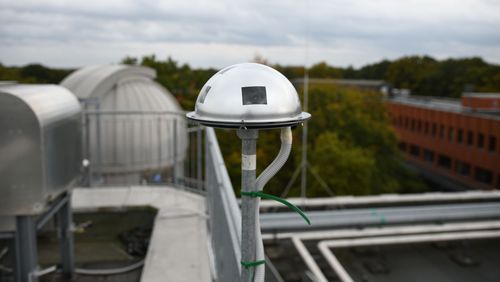 Die Kamera ist ein kuppelförmiges Metallteilt mit einigen Öffnungen. Sie befindet sich auf einer Stange auf dem Dach eines Gebäudes, im Hintergrund die Kuppel des Observatoriums.
