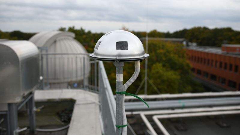 Die Kamera ist ein kuppelförmiges Metallteilt mit einigen Öffnungen. Sie befindet sich auf einer Stange auf dem Dach eines Gebäudes, im Hintergrund die Kuppel des Observatoriums.