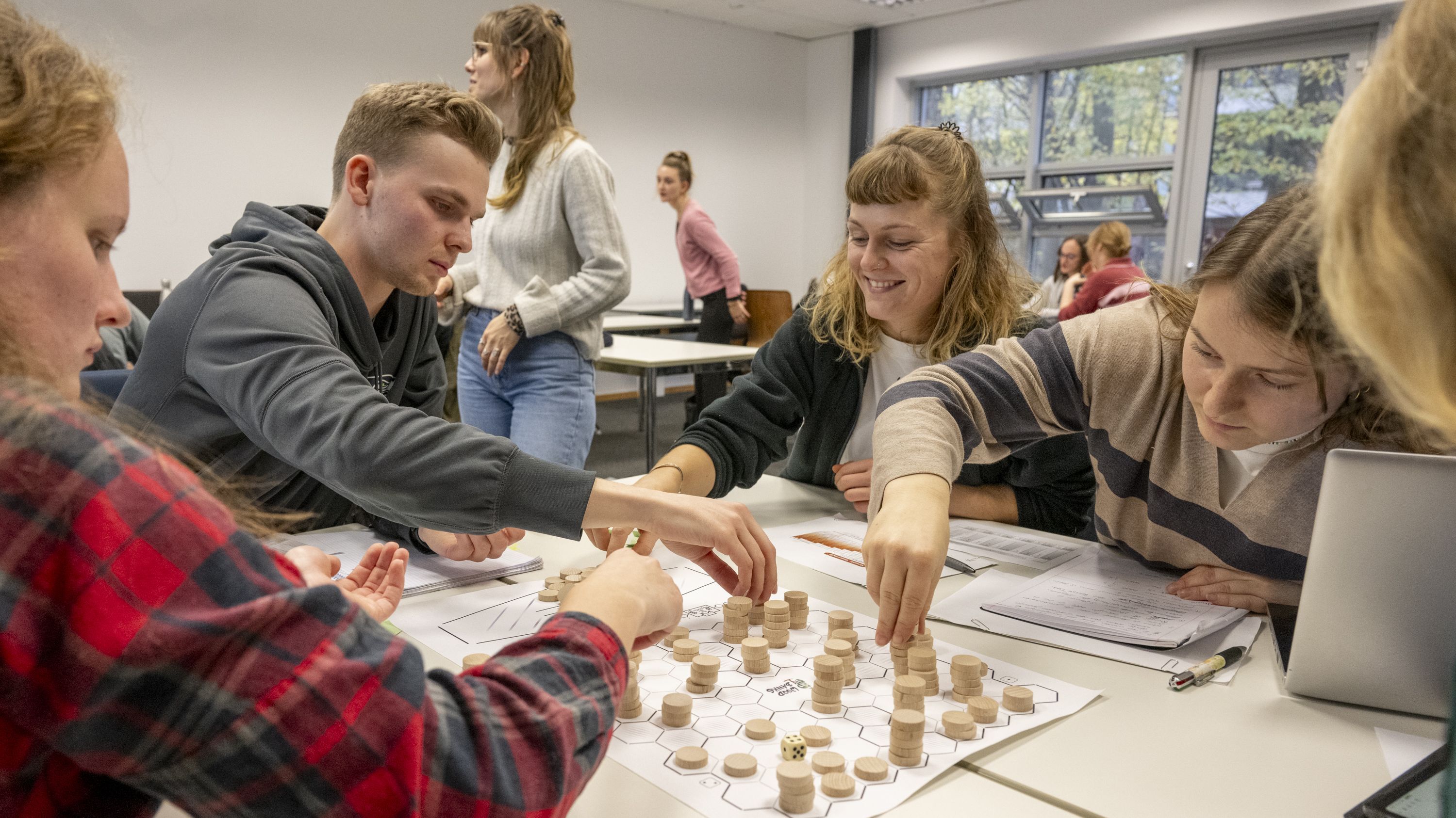 Das Bild zeigt eine Gruppe von Studierenden, die das Spiel "Woodbanks" spielen. In der Tischmitte liegt das Spielfeld, darauf stehen kleine hölzerne Chips, die von den Spielenden bewegt werden. Auch ein Würfel liegt auf dem Tisch.