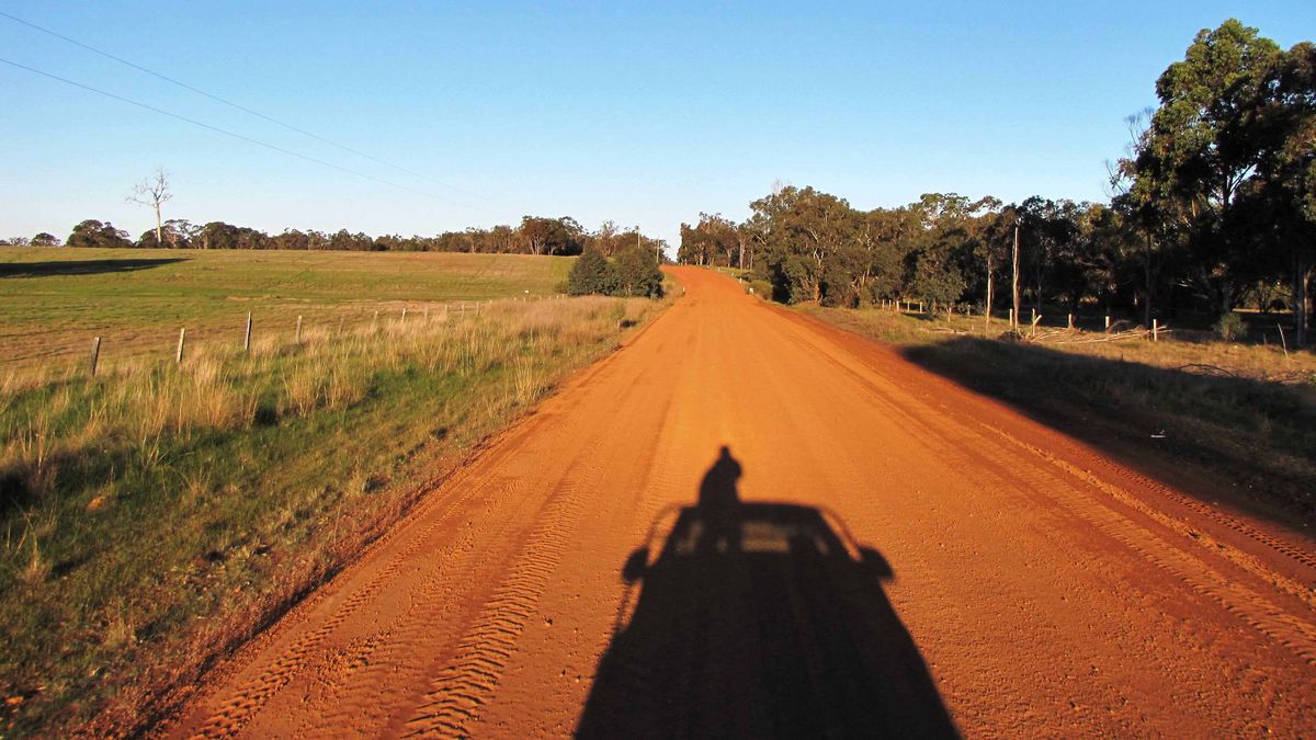 Eine lange Landstraße durch eine hügelige Landschaft mit grünen Feldern und Bäumen. Ein Schatten einer Person im Fahrzeug.