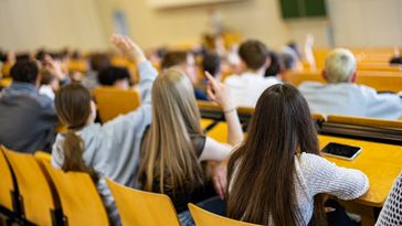 People are sitting on wooden chairs in the lecture hall, pointing their fingers in the air or raising their hands.