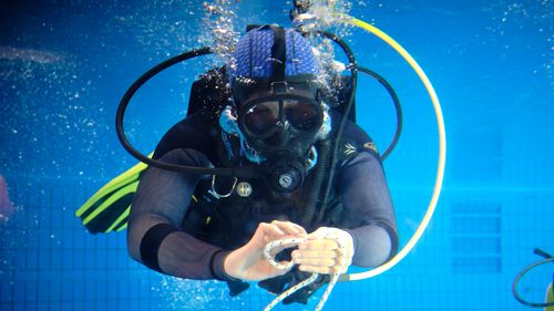 Underwater picture. A person in a diving suit with mask, oxygen tanks and fins knots a thick cord.