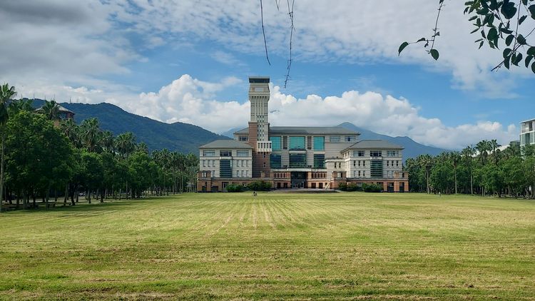 The main building of the university behind a mown meadow, with trees to the side and wooded mountains in the background. 