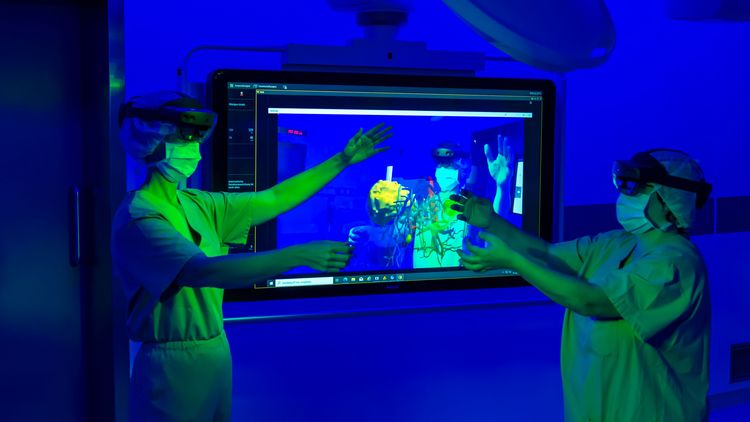 Two people in surgical gowns with data glasses move their arms so that they move the virtual organs. In the background, a large screen shows what they can see in the glasses.