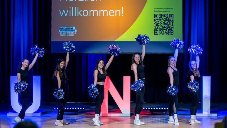 Cheerleaders wave their pom-poms up and down. Behind them, a screen displays the word: Welcome.