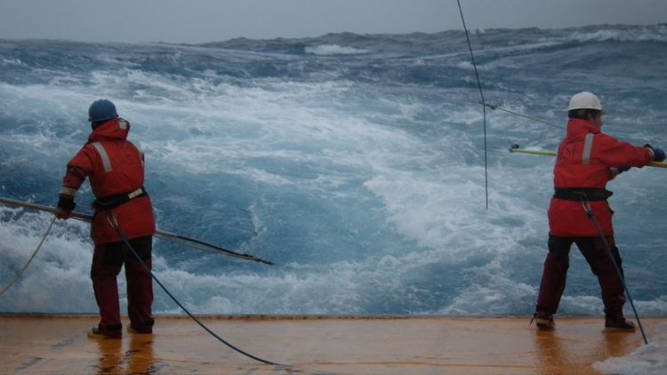 Two men on board the research vessel with poles in their hand to retrieve coral samples. The churning sea is in the background.