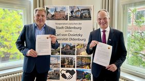 Zwei Männer stehen vor einem Aufsteller und halten ein Blatt Papier in die Kamera. 