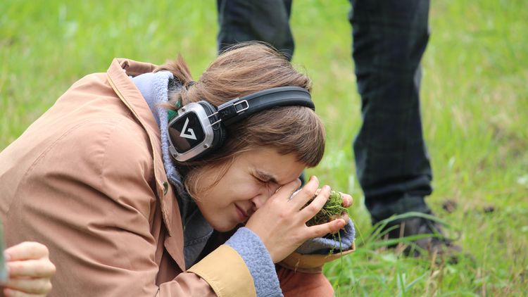 Eine Frau ist von der Seite zu sehen. Sie hockt auf einer Wiese und hält sich ein Büschel Gras oder ein Stück Wiese direkt vor ein Auge. AUßerdem trägt sie Kopfhörer.