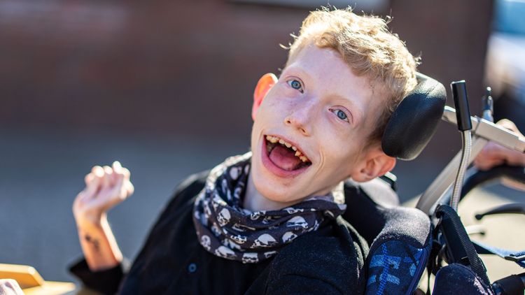 Das Bild zeigt einen jungen Mann, der unter einer schweren Beeinträchtigung leidet. Er sitzt in einem Rollstuhl. Er lacht sehr herzlich in die Kamera. Im Hintergrund ist ein Haus zu erkennen.