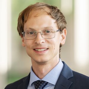 The picture shows Peter Eppinger. He is standing in a corridor in a university building and smiles at the camera. He is wearing a badge with the university logo on his lapel.