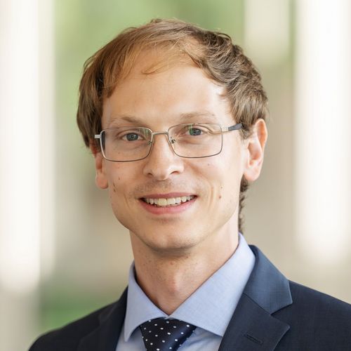 Das Bild zeigt Peter Eppinger. Er steht auf einem Flur in der Uni und lächelt in die Kamera. Er trägt einen Anstecker mit Unilogo am Revers.