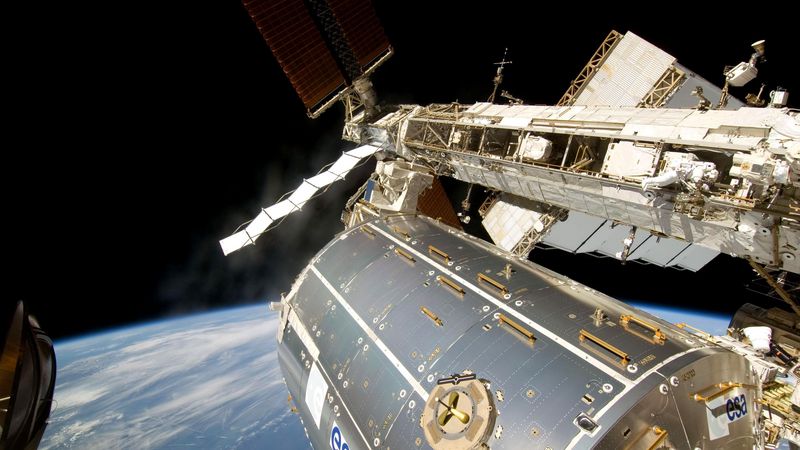 Das zylinderförmige Forschungsmodul Columbus schwebt im Weltraum, im Hintergrund ist die Erde zu sehen. 