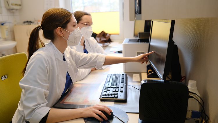 Antonia und Alexandra sitzen im Arztzimmer gemeinsam vor einem PC und besprechen sich.
