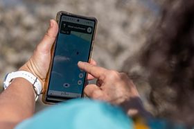 Blick von hinten über die Schulter einer Frau, die ein Smartphone in der Hand hält. Auf dem Bildschirm ist Google Maps zu sehen, die Position des Sensors ist als rotes Fähnchen zu sehen.