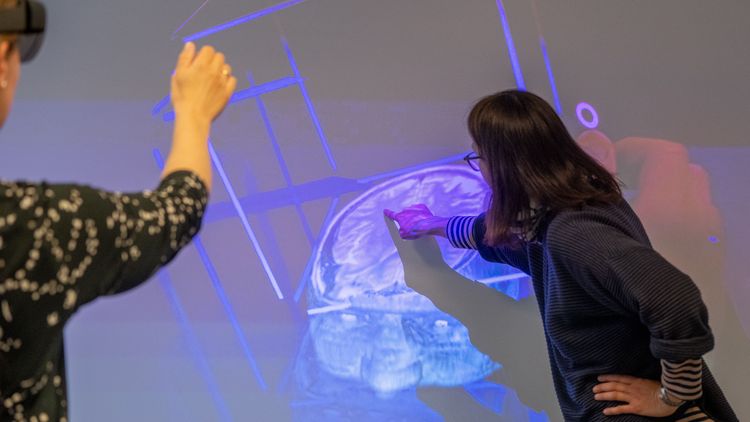 Eine Frau mit AR-Brille macht eine Geste, eine andere Frau zeigt auf die Projektion eines Gehirns auf einer Leinwand.