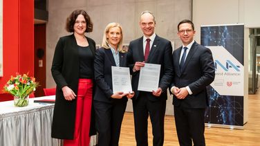 Das Bild zeigt die vier Personen. Sie stehen vor dem Logo der Northwest Alliance und lächeln in die Kamera. Jutta Günther und Ralph Bruder halten das unterzeichnete Memorandum of Understanding in die Kamera. Links von ihnen befindet sich ein Tisch, auf dem eine Vase mit bunten Blumen steht.