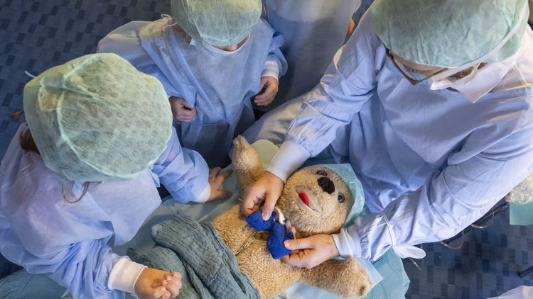 Blick von oben: Ein Teddy liegt auf einem improvisierten OP-Tisch. Sein Reißverschluss-Bauch ist geöffnet. Eine Frau hält ein blaues Plüschteil in den Händen, das die Lunge des Teddys darstellt. Zwei Kinder in OP-Kleidung assistieren.