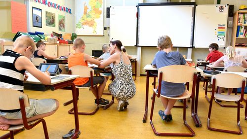 Schülerinnen und Schüler in einem Klassenzimmer. Die Kinder sitzen auf braunen Stühlen und wenden der Kamera dem Rücken zu. Neben einem Schüler kniet die Lehrerin, um etwas zu erklären.