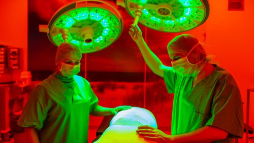 Two doctors in surgical clothing stand in front of the operating table and move the large, round operating lamp by hand.