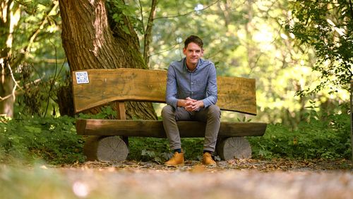 Dominik Gautier sits on a wooden bench in a forest and looks into the camera.