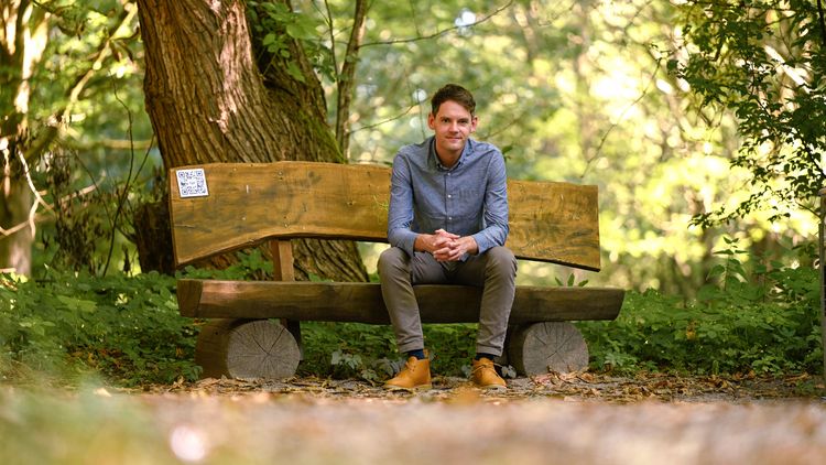 Dominik Gautier sitzt auf einer Holzbank in einem Wald und blickt in die Kamera.