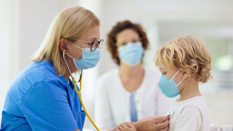 Ärztin mit Maske untersucht kleinen Jungen, ebenfalls mit Maske, mit dem Stethoskop.