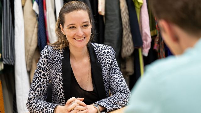 The photo shows Klara von Lindern in the rooms of the Everyday Textile Culture Collection. In the background is an open closet in which numerous different and colorful items of clothing are hanging, mainly jackets and coats. Klara von Lindern is sitting at a table in the center. She has her hands folded in front of her and smiles towards the interviewer. He can be seen in the right foreground.