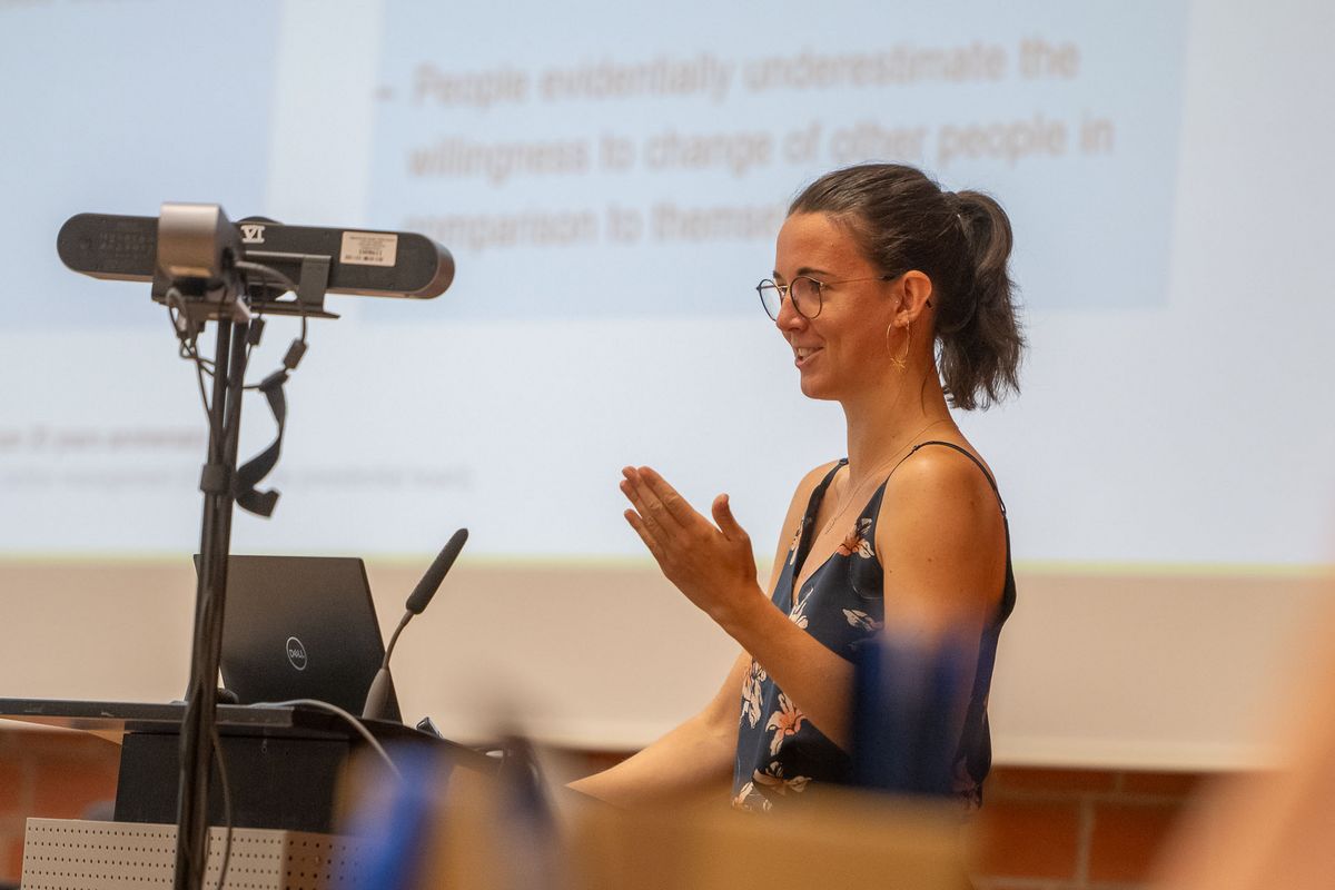 Eine Frau mit dunklem Haar und Brille steht vor einer Projektion und spricht. Sie trägt ein blaues Top mit Blumenmustern und lächelt, während sie mit ihren Händen gestikuliert. Vor ihr steht ein Laptop und ein Mikrofon auf einem Pult. Der Hintergrund ist verschwommen, aber man kann erkennen, dass sie sich in einem Konferenzraum befindet. Das Bild vermittelt den Eindruck einer Präsentation oder eines Vortrags während des Symposiums.