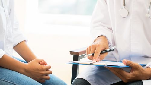 Eine junge Frau sitzt neben einem Arzt, der etwas auf einem Klemmbrett zeigt. Die Hände befinden sich in der Mitte des Bildes.