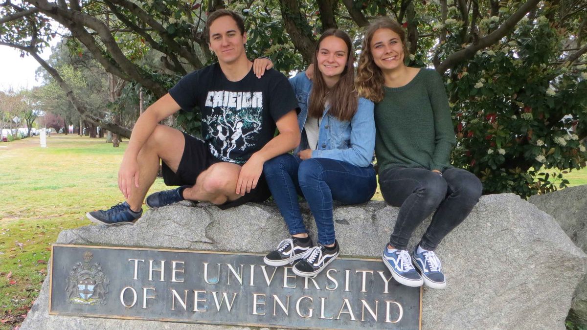 Three young people are sitting on a rock and smiling. In front of them is a bronze plaque with the coat of arms and the words "The University of New England".