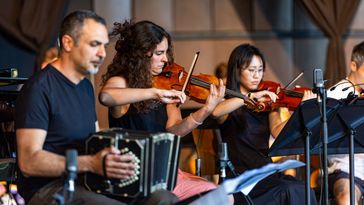 Die Musiker des Ensembles sind gerade bei der Probe. Zu sehen sind ein Mann, der Bandoneon spielt sowie zwei Violinistinnen. Alle sitzen hinter Notenpulten. DAs Bandoneon sieht ähnlich aus wie ein kleines Akkordeon.