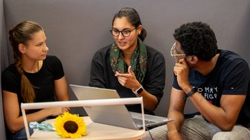 Three people sit next to each other in an alcove and discuss, with a laptop open on the mobile table in front of them.
