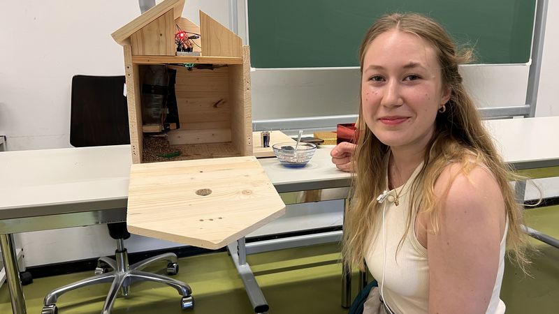 Eine Studentin blickt in die Kamera. Neben ihr ist ein Vogelhaus aus Holz zu sehen, die Front ist aufgeklappt. Im Haus befindet sich ein Fallrohr, darunter liegt ein kleiner Haufen Vogelfutter. 