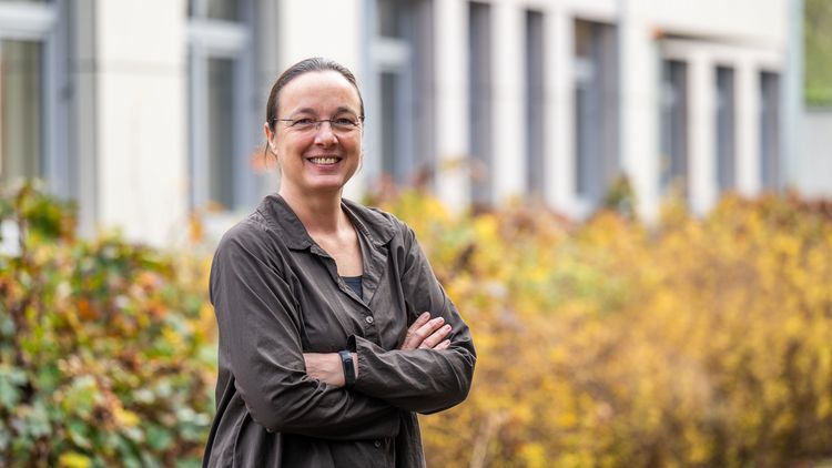 Eine Frau steht vor einer herbstlich anmutenden Hecke, dahinter ist ein Gebäude zu erkennen.