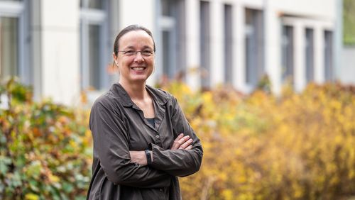 Eine Frau steht vor einer herbstlich anmutenden Hecke, dahinter ist ein Gebäude zu erkennen.