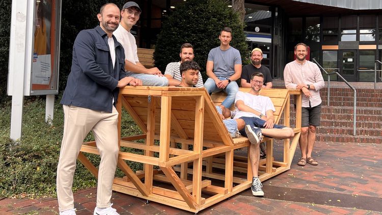 Menschen stehen um eine Sitzbank aus Holz.