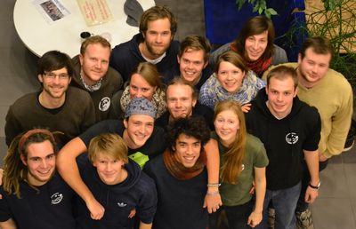 The photo shows the Environmental Sciences Student Council for winter & summer semester 2012/13. 15 students can be seen, all in their early to late 20s, all wearing colourful jumpers, jackets or T-shirts with the Student Council logo.  They are standing in several rows. The photo was taken in the ICBM and from far above the people's heads. A grey tiled floor and two tables can be seen in the background.