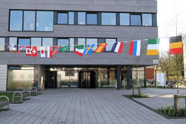 Das SSC (StudierendenServiceCenter) der Universität Oldenburg, ein modernes Gebäude mit grauen Betonplatten und großen Glasflächen. Vor dem Gebäude hängen Flaggen verschiedener Länder.