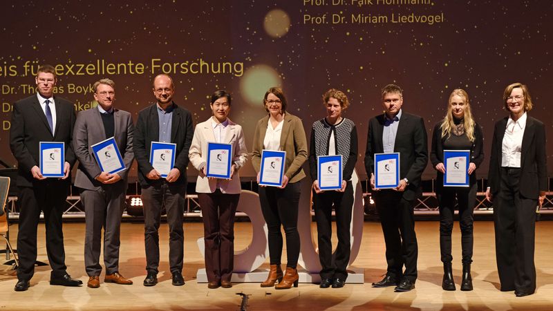 Acht Preisträger*innen halten ihre Urkunden in der Hand, daneben steht UGO-Vorsitzende Wiebke Schneidewind.