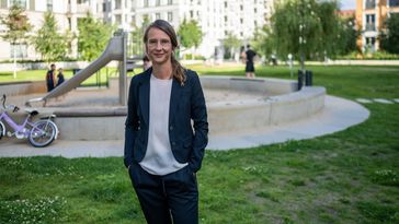 Das Bild zeigt Gundula Zoch. Sie steht am Rande eines Spielplatzes in einer Stadt. Hinter ihr spielen Kinder, eine Rutsche steht im Sand. Weitere Menschen machen Sport. Zoch lächelt in die Kamera und hält ihre Hände in den Hosentaschen.