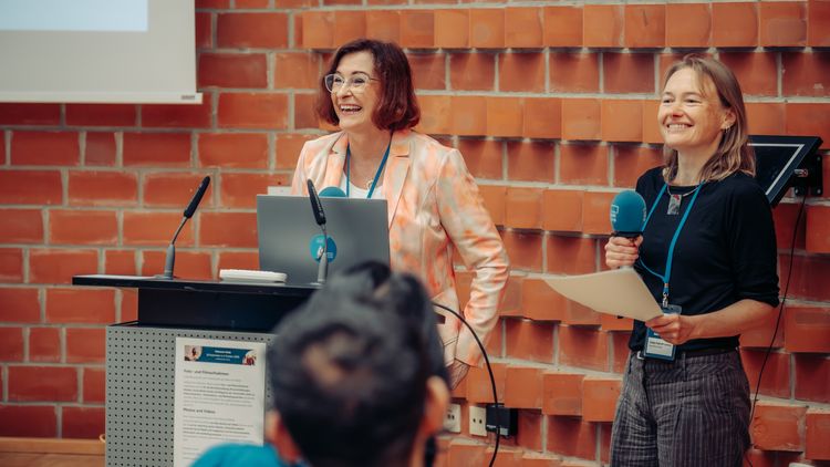 Andrea Strübind, Vizepräsidentin für Studium und Lehre, und Katja Kaboth vom Welcome Team für internationale Studierende bei der Begrüßungsveranstaltung. Frau Strübind steht an einem Schreibtisch mit Laptop und Mikrofon, Frau Kaboth hält ein blaues Mikrofon und ein Blatt Papier in der Hand. Beide stehen vor einer roten Backsteinwand.