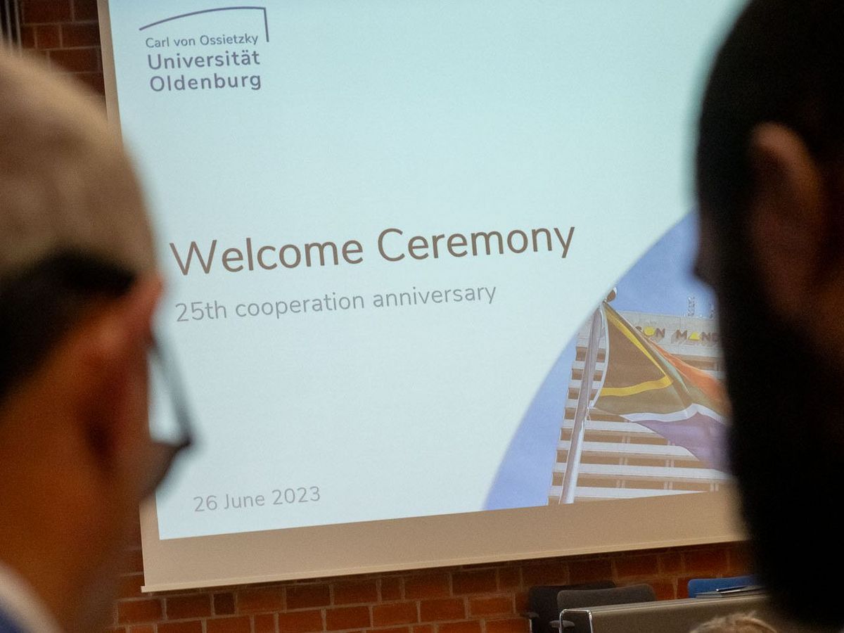 Das Bild zeigt eine Projektion auf eine Leinwand. Auf der Leinwand steht in großen Buchstaben "Welcome Ceremony" und darunter "25th cooperation anniversary". Darunter ist das Datum "26 June 2023" angegeben. Oben auf der Leinwand ist das Logo der Carl von Ossietzky Universität Oldenburg zu sehen. Im Vordergrund sind die Köpfe von Personen zu sehen, die der Leinwand zugewandt sind. 