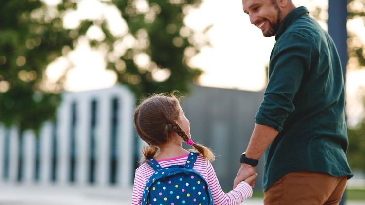 Papa und Kind mit Rucksack Hand in Hand auf dem Campus, Papa bringt Kind zum Kindergarten
