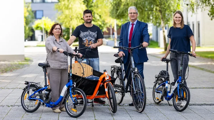 Uni-Mitarbeitende mit den vier zentralen Campus-Rädern, das sind zwei Klappräder, ein Stadtrad  mit Gepäckträger und niedrigem Einstieg sowie ein Lastenrad mit großer offener Ladebox vorne und einer Transportbox auf dem Gepäckträger.