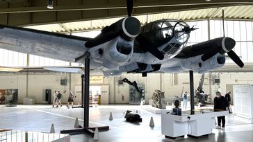 Das Bild zeigt ein Modell eines Bombers, das in einer großen Museumshalle steht. Der Schacht des Flugzeugs ist geöffnet, einige Bomben fallen heraus. Im Hintergrund ist das im Text erwähnte Friesfragment zu erkennen. In der Halle stehen einige Besucher und betrachten die Ausstellung.