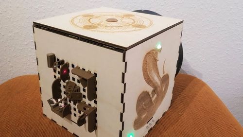 Edge view on a wooden cube. On one side, there is an image of a cobra, on the other a sun wheel, on the third plastic models of Aztec buildings. Not to be seen is the fourth side with a vault opening.