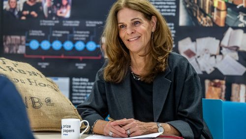 The picture shows Dagmar Freist talking to the interviewer. She is smiling at him. Next to her is a cup with the words "Prize Papers Project" written on it. Next to it lies a jute bag. In the background are several posters with information about the Prize Papers project.