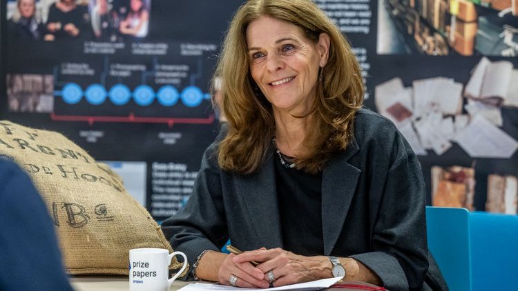 The picture shows Dagmar Freist talking to the interviewer. She is smiling at him. Next to her is a cup with the words "Prize Papers Project" written on it. Next to it lies a jute bag. In the background are several posters with information about the Prize Papers project.