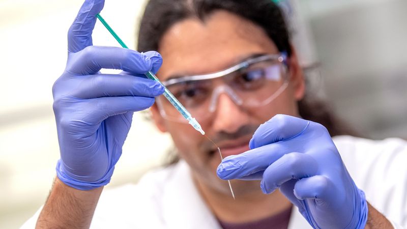 Ein Wissenschaftler hat eine schmale Pipette in der Hand, mit der er einen haarfeinen Draht in der Druckdüse aus Glas platziert. 